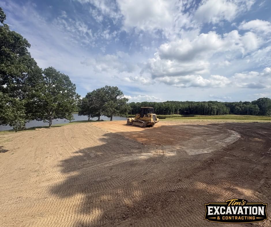 Bulldozer grading a dirt lot beside trees under a partly cloudy sky