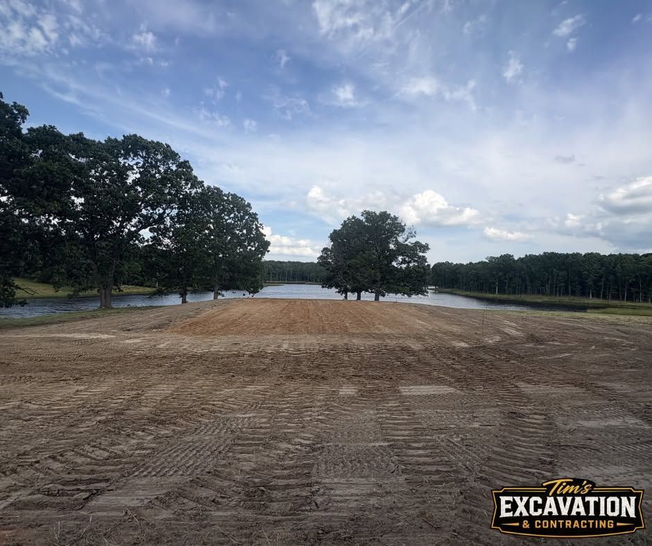 Cleared dirt lot beside a lake under a cloudy sky, with trees lining both sides and a distant bridge.