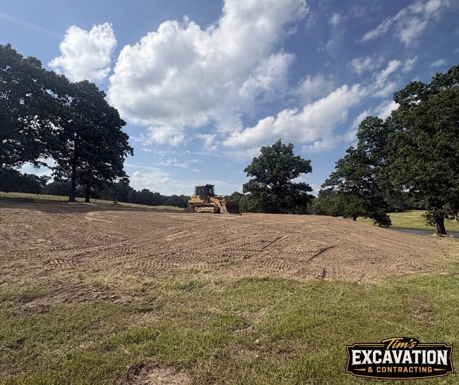 Bulldozer grading a wide dirt field under a partly cloudy sky, with trees around the edge