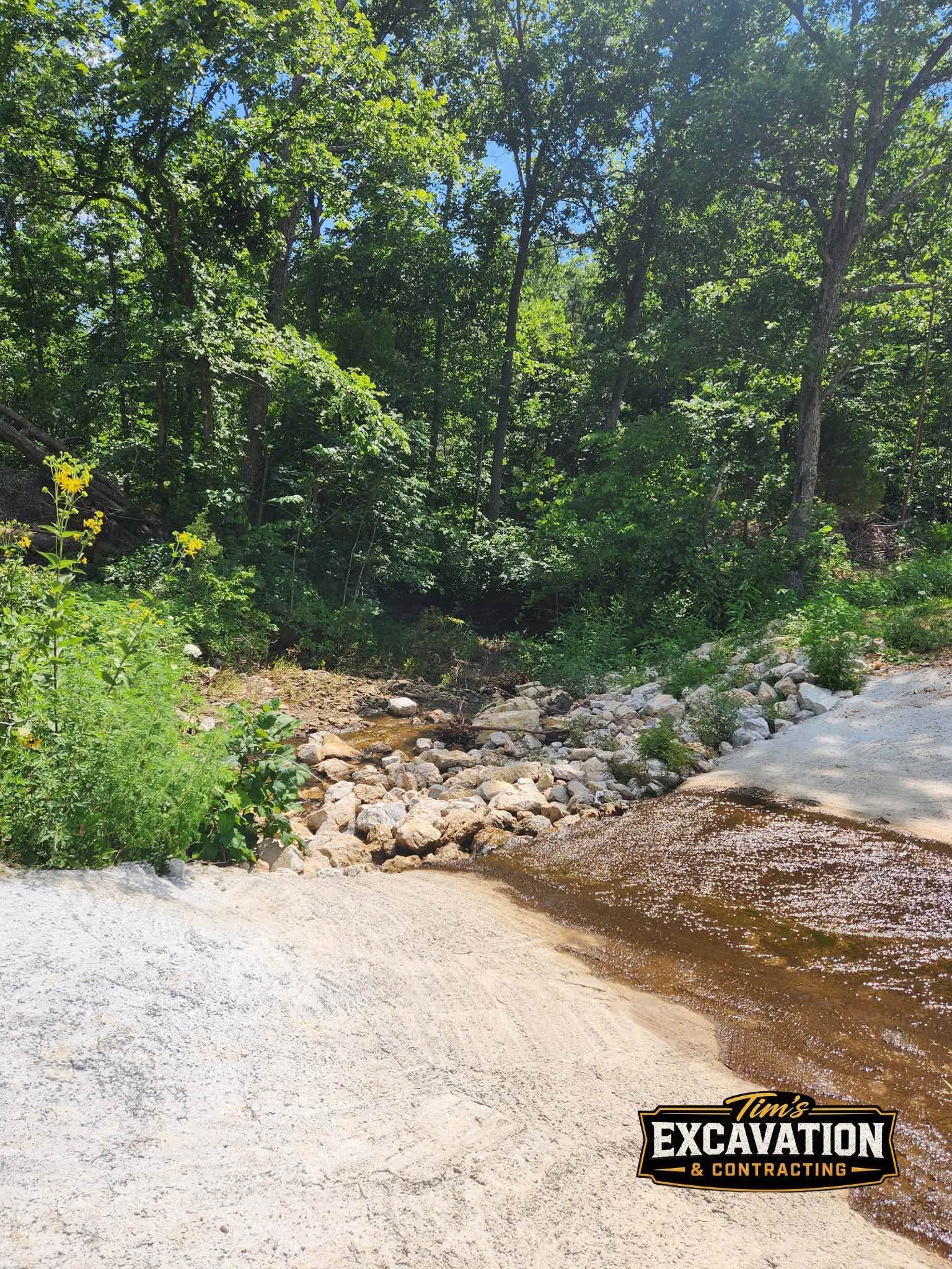 Shallow stream over rocks with gravel bank and dense green trees in sunlight