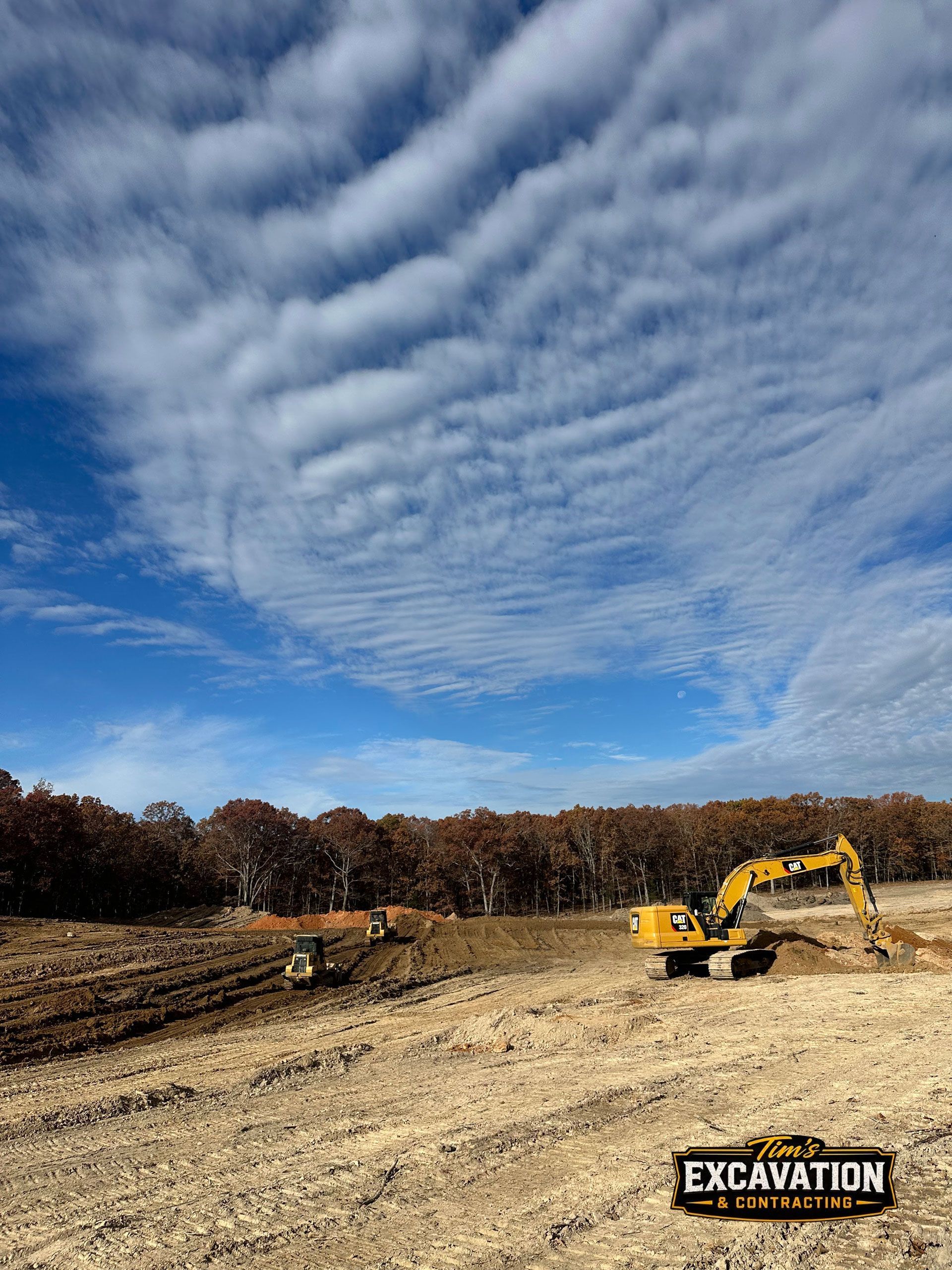 Excavator working on a dirt site under a cloudy blue sky, with trees lining the background