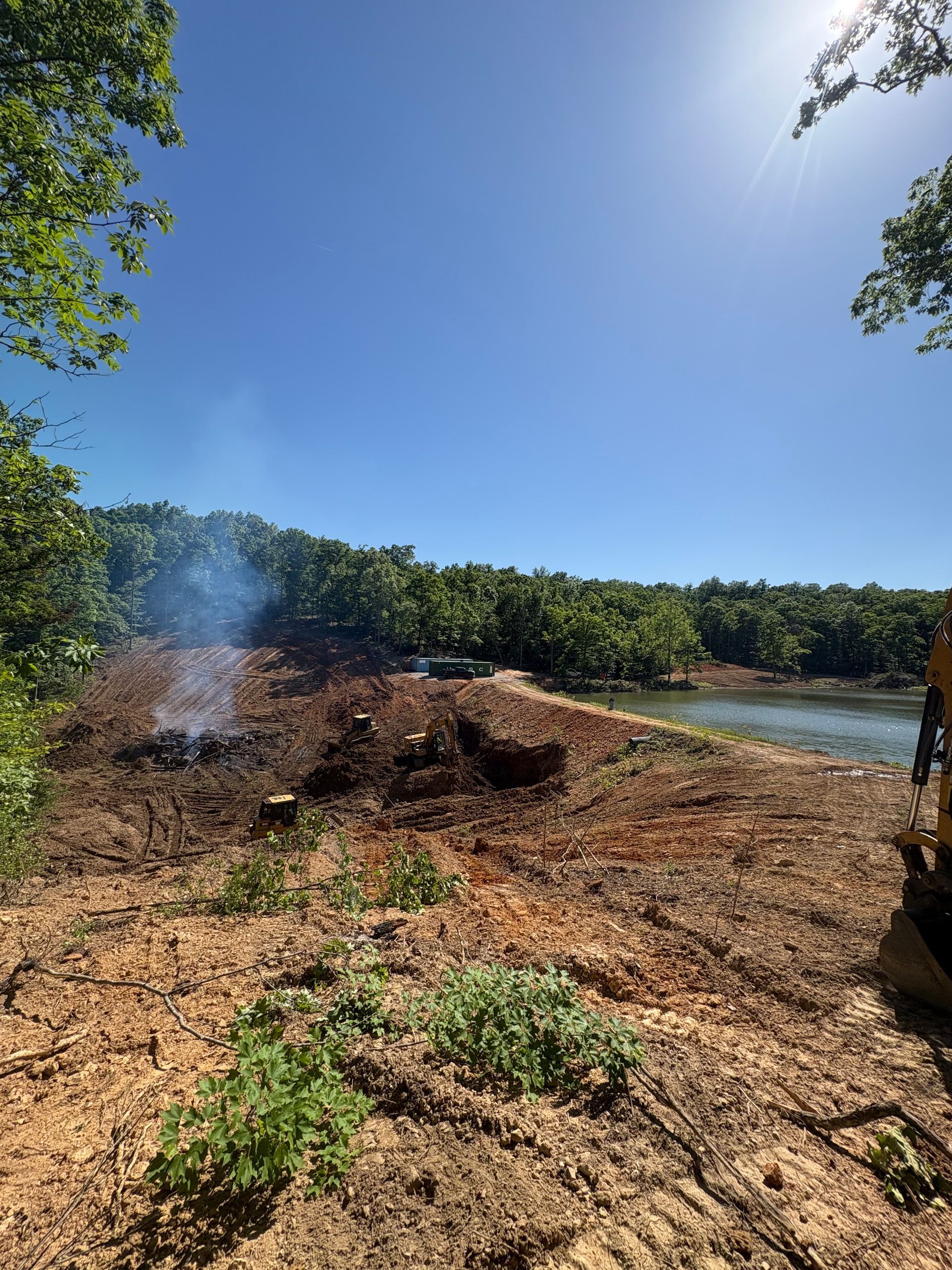 Brush pile burning near a wooded riverbank under a bright blue sky
