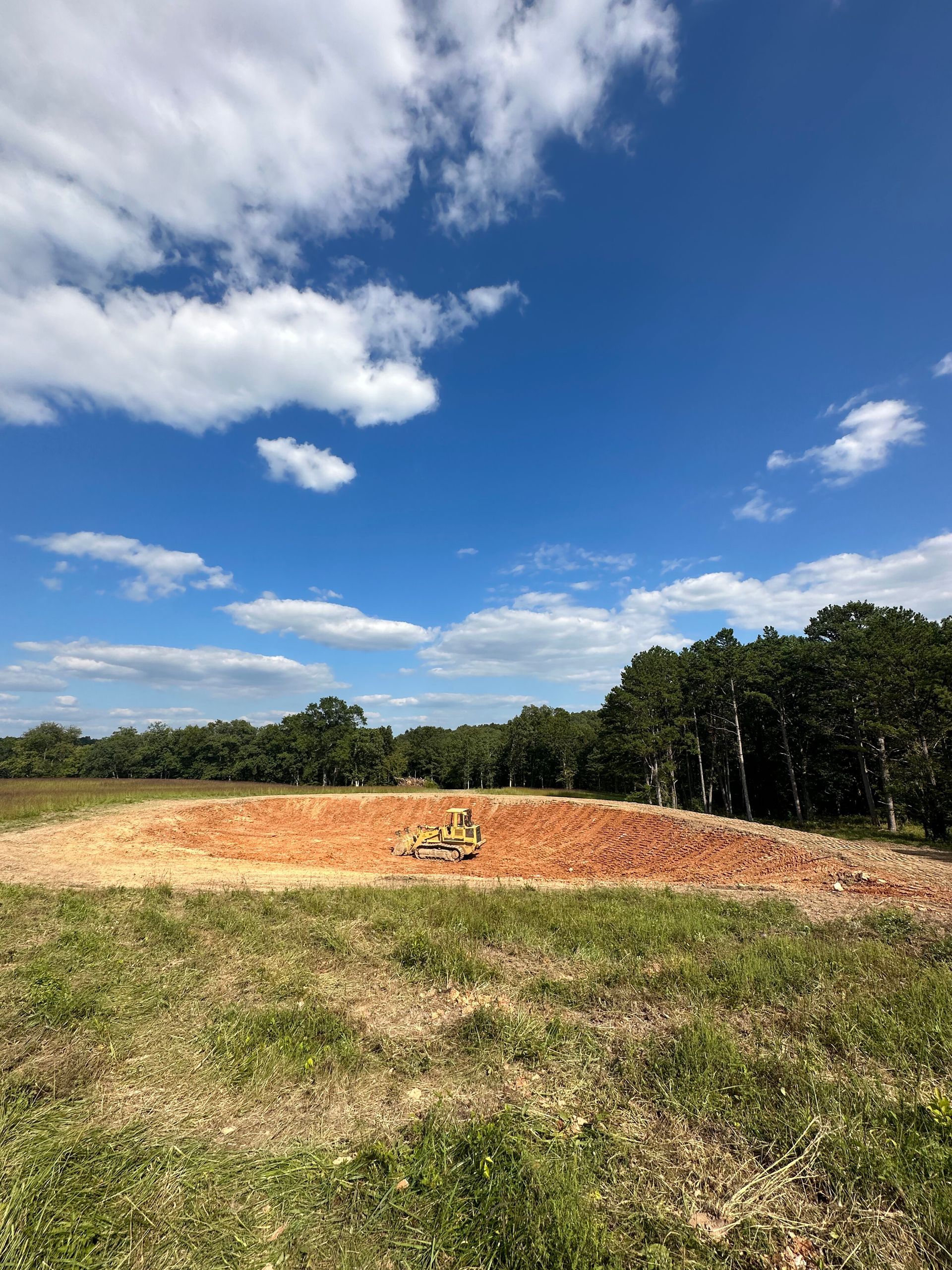 Grassy field with a small dirt mound under a bright blue sky with scattered clouds.