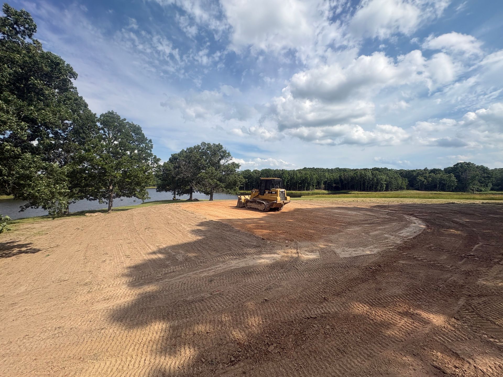 Bulldozer grading a dirt field under a partly cloudy sky, with trees and a river in the background