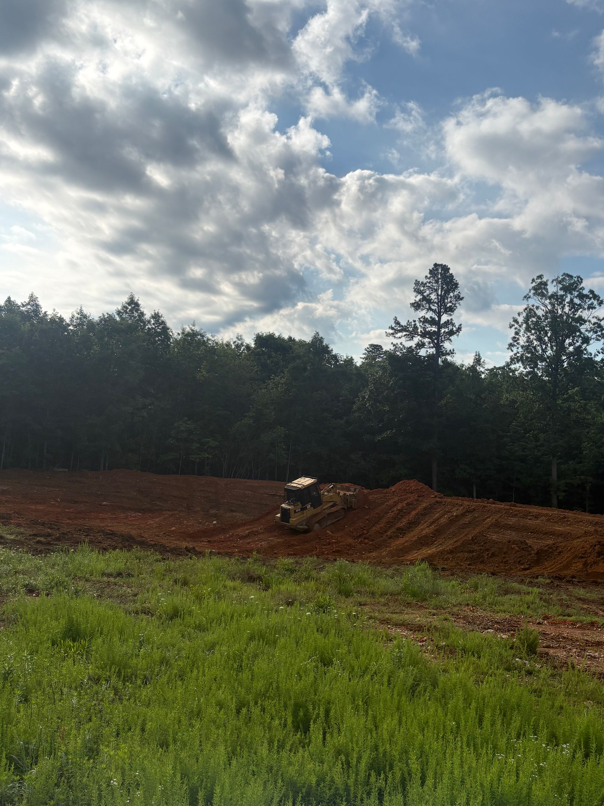 Bulldozer grading red dirt field beside a grassy foreground and tree line under cloudy sky