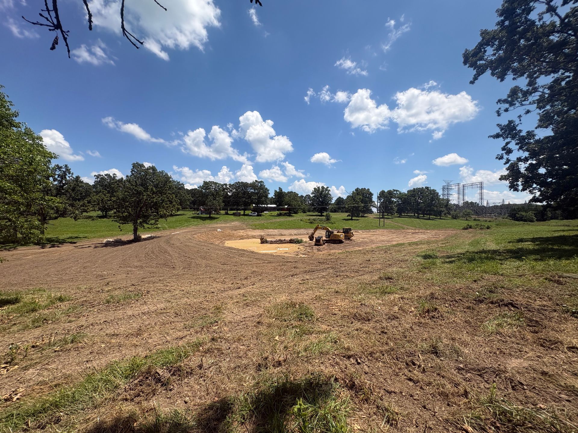 Open grassy field under a blue sky with scattered clouds, trees lining the horizon, and a few logs in the center