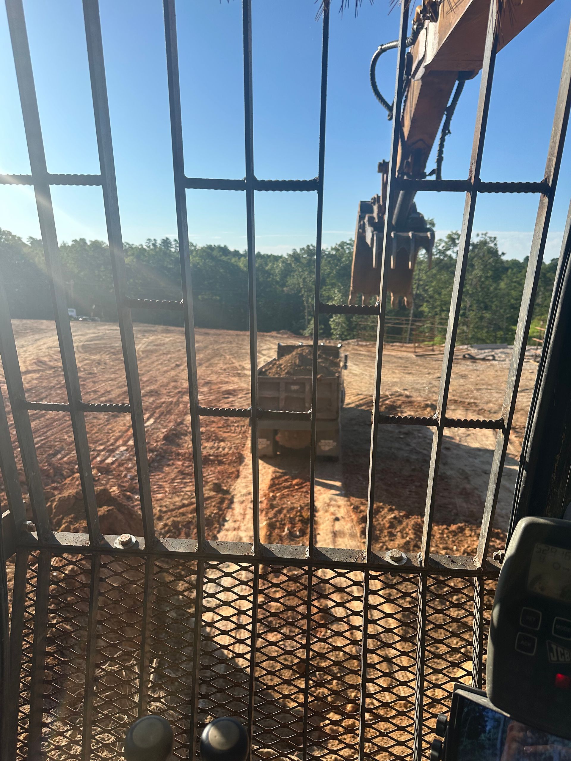 View through a metal gate to a dirt lot with a wooden crate and excavator arm under a bright blue sky