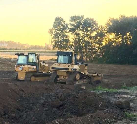 Gravel lot under stormy clouds with a fenced dirt field and trees in the background, excavations logo at bottom right