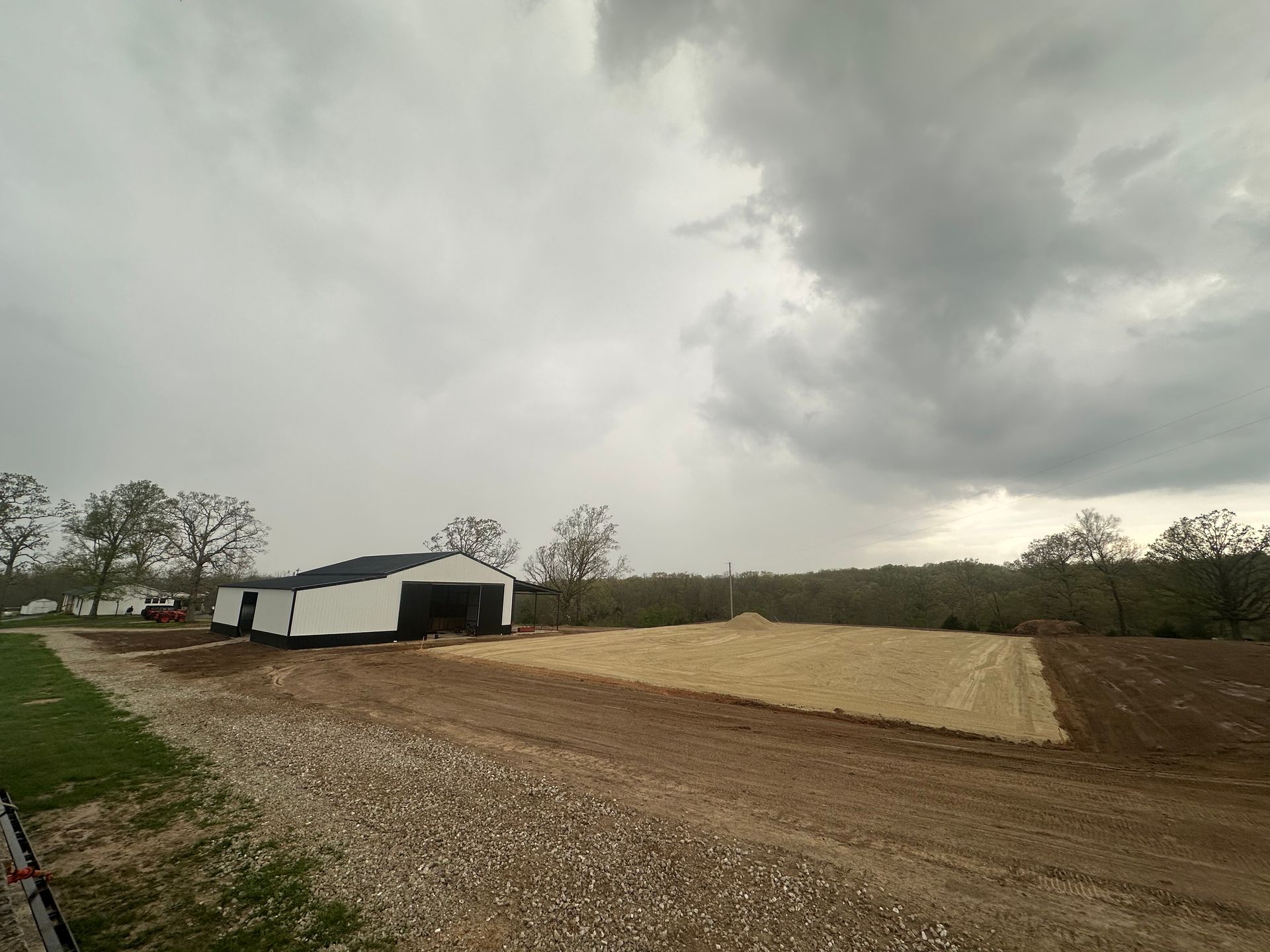 Rural farmyard with white barns, gravel path, and freshly plowed field under a cloudy sky