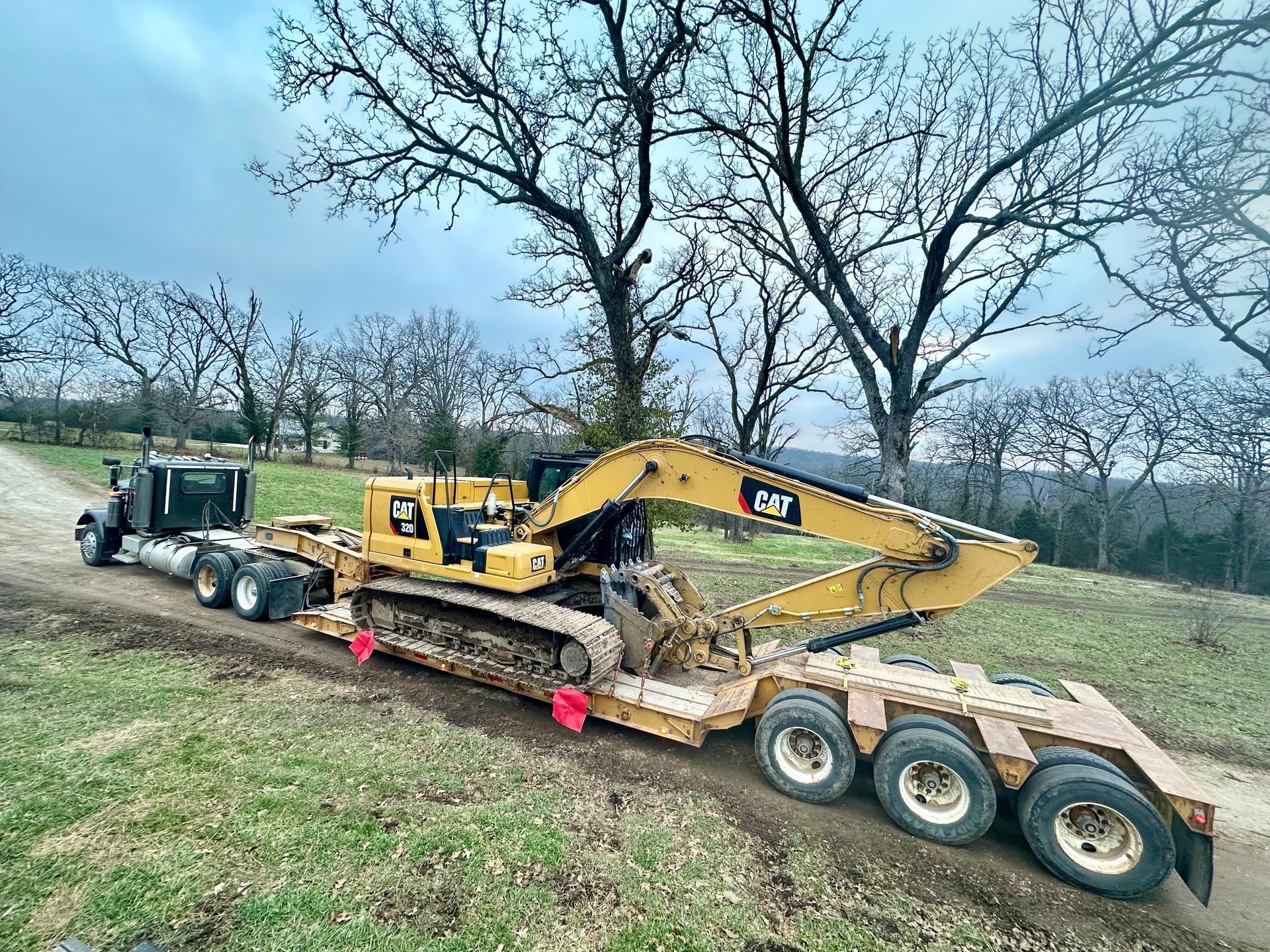 Yellow excavator loaded on a lowboy trailer being towed along a rural dirt road