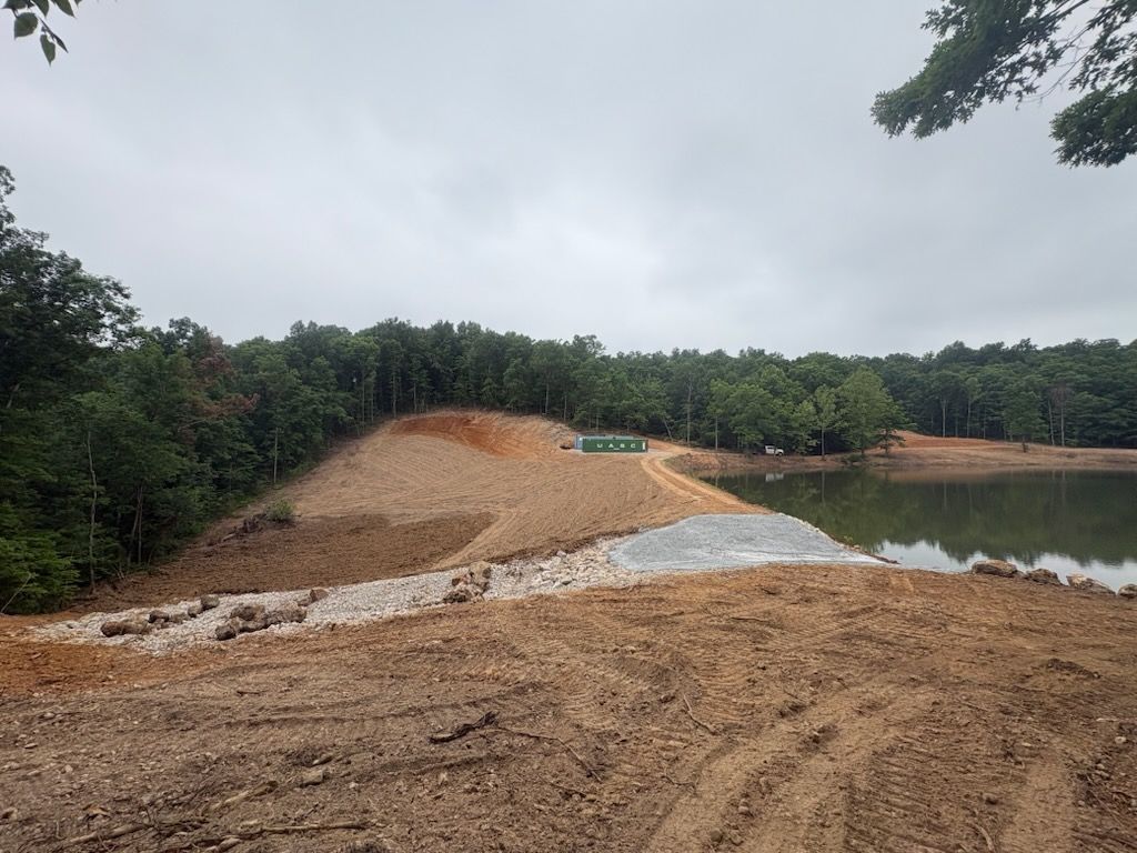 Gravel mound beside a calm pond, with trees lining the shore under an overcast sky.