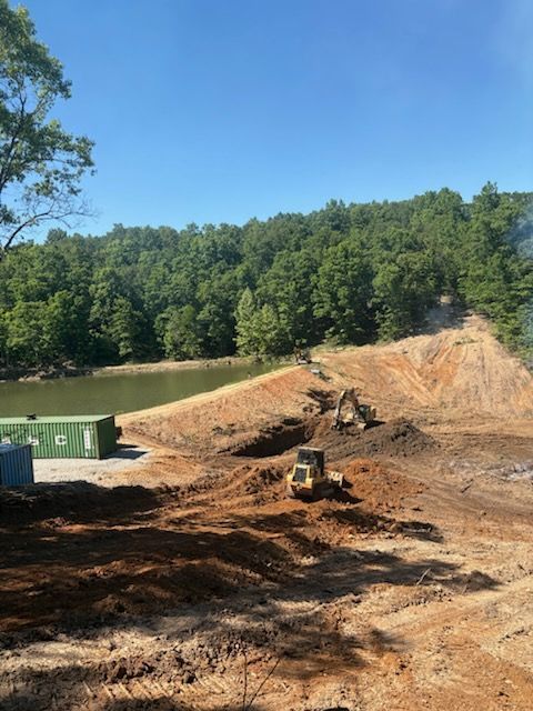 Construction site with earth-moving equipment beside a muddy pond and green dumpster, backed by trees under blue sky