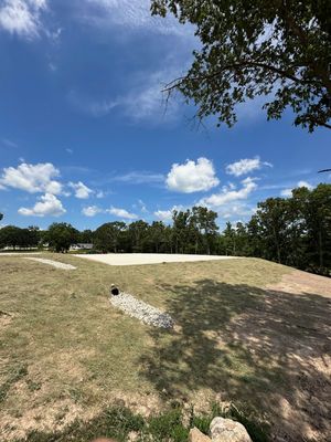 Sunny grassy hill with a small concrete pipe, trees, and a blue sky with scattered clouds