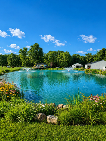 Bright blue pond with fountains, green grass, flowers, and trees under a sunny sky