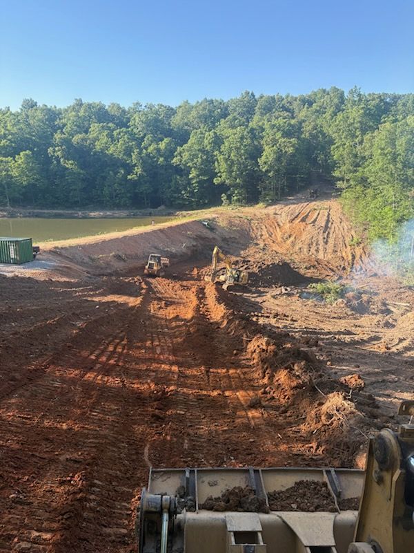 Excavator working on a dirt road beside a pond, with wooded hills in the background.