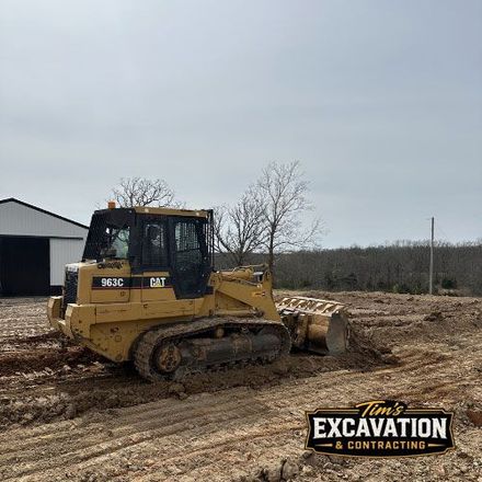 Yellow CAT bulldozer grading muddy dirt lot near a barn under a gray sky