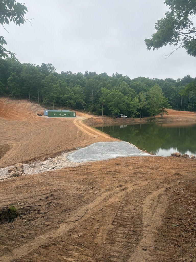Road construction site beside a muddy pond, with dirt piles, a gravel path, and trees under cloudy skies