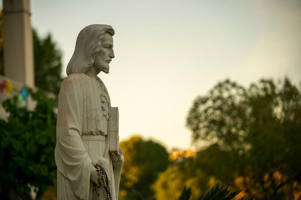White statue of a robed figure outdoors at sunset, with trees and a building in the background