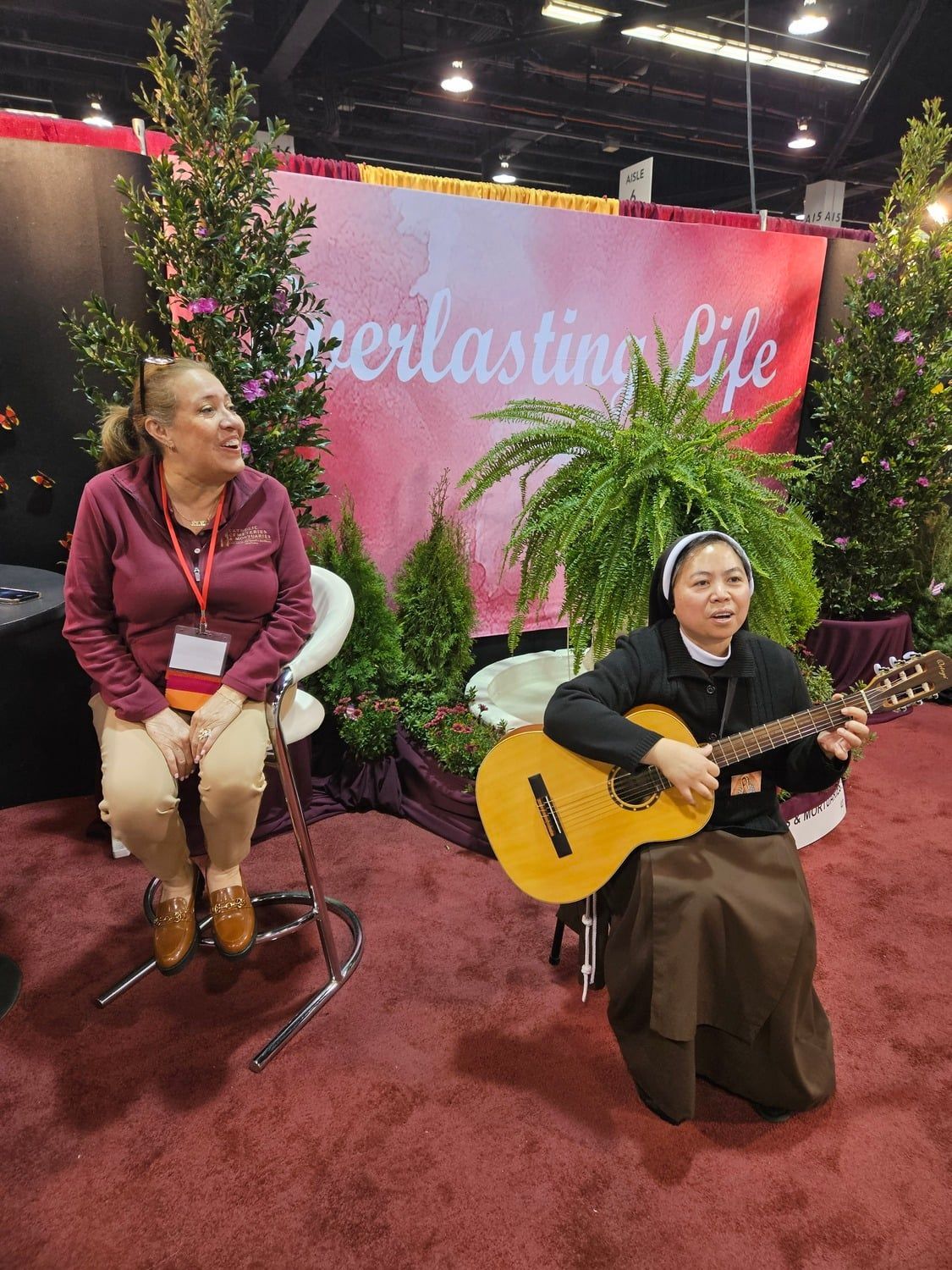 Two women at an indoor booth; one plays guitar while the other sits beside plants and a pink “Everlasting Life” backdrop.