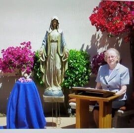 Woman seated at a lectern beside a statue, with flower arrangements on either side