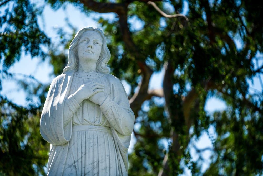 White statue of a robed figure with hands crossed, framed by sunlit green trees and blue sky