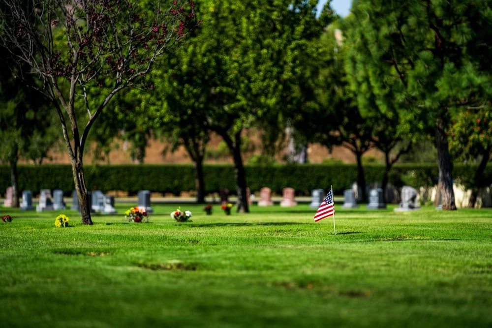 Grassy cemetery with headstones, trees, and a small American flag marking a grave