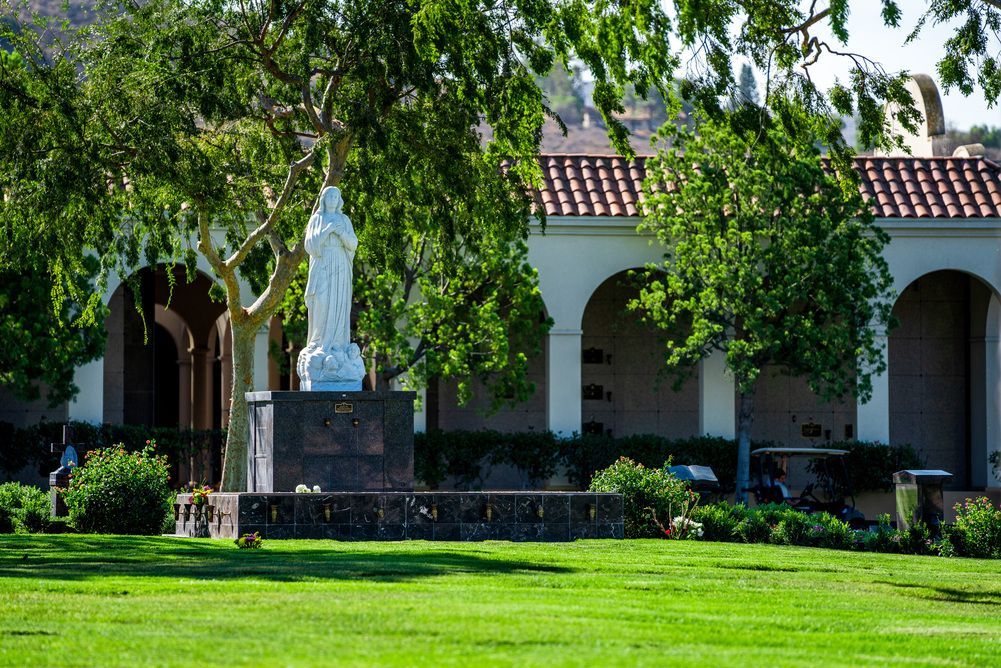 Statue fountain in front of a white arched building, shaded by trees with a green lawn in the foreground