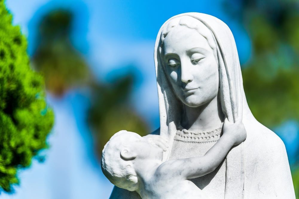 White statue of a robed figure with a bird on its shoulder, set against a blue sky and green trees.