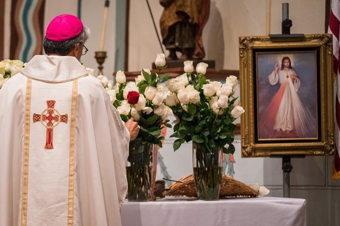 Priest in white vestments and pink skullcap stands at altar with flowers beside framed image of Jesus' Divine Mercy