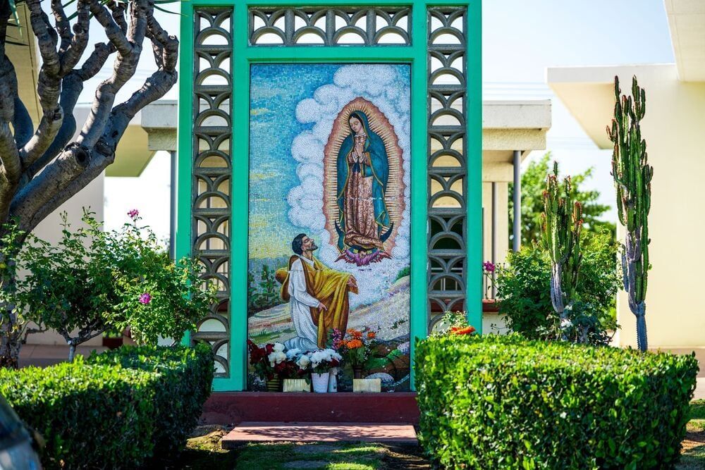 Outdoor shrine mural of the Virgin of Guadalupe framed by turquoise columns and garden hedges
