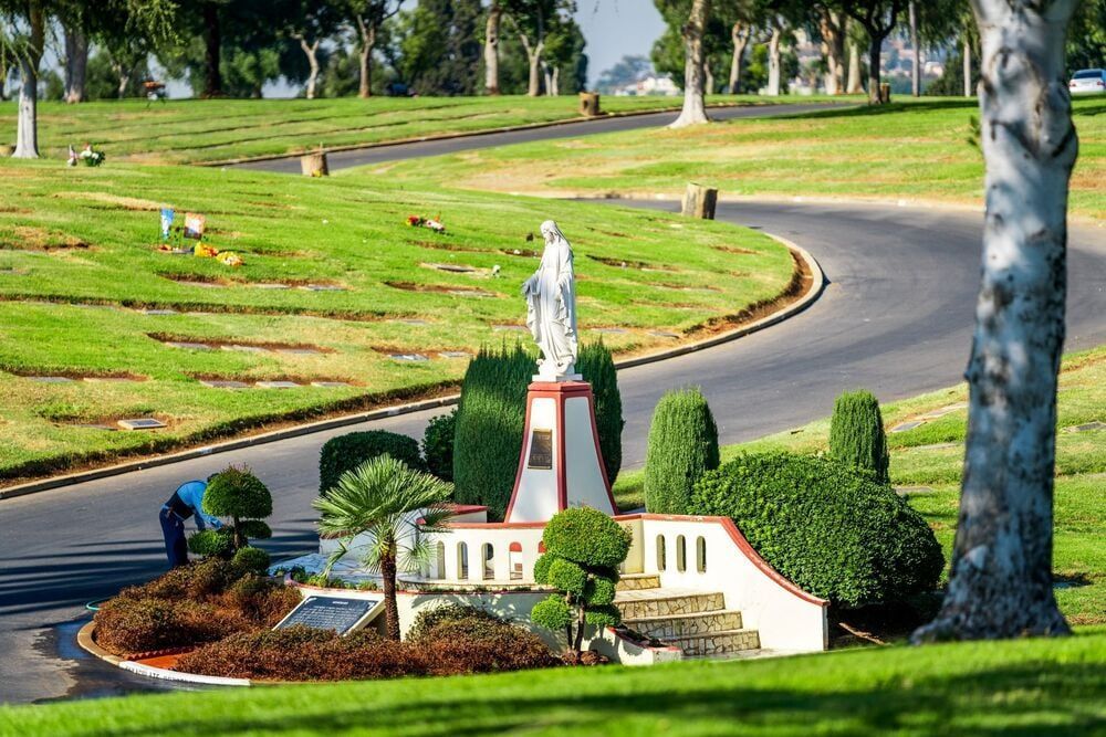 Miniature roadside shrine in a landscaped park beside a winding road and green hillside.