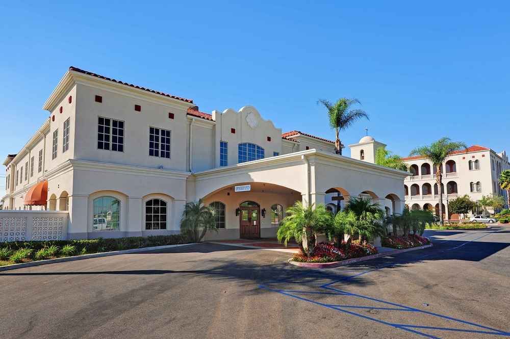 Mediterranean-style hotel entrance with palm trees and a covered driveway under a blue sky