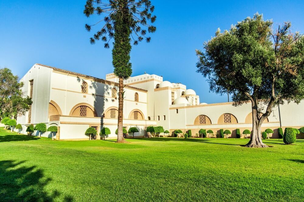 Cream-colored villa with arches and dome, set behind a green lawn and trees under a bright blue sky