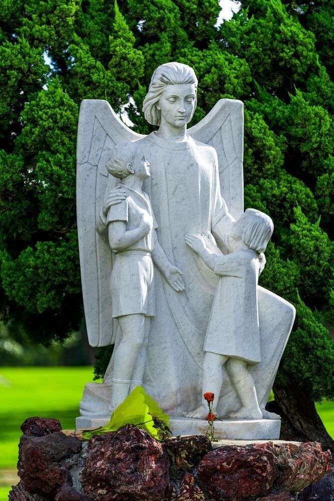 White stone angel statue with two children in a green garden, flowers at the base