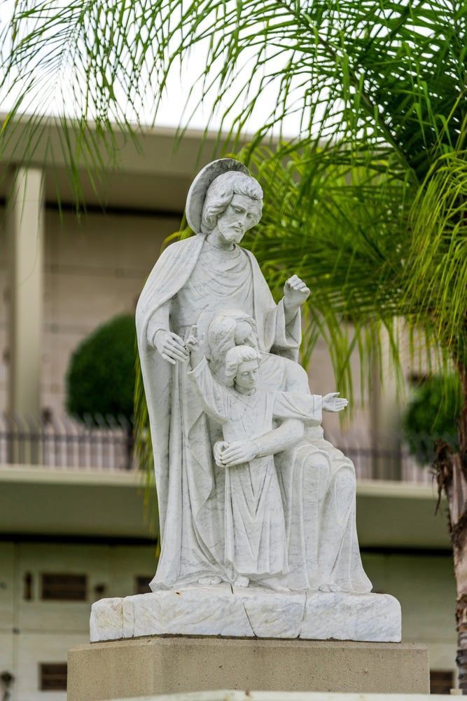 White religious statue of a robed figure holding a child, set among palm trees outdoors