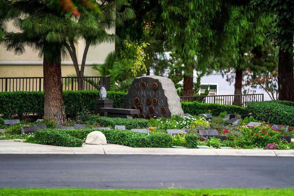 Landscaped park with trees, flowers, a stone fountain, and a road in front.