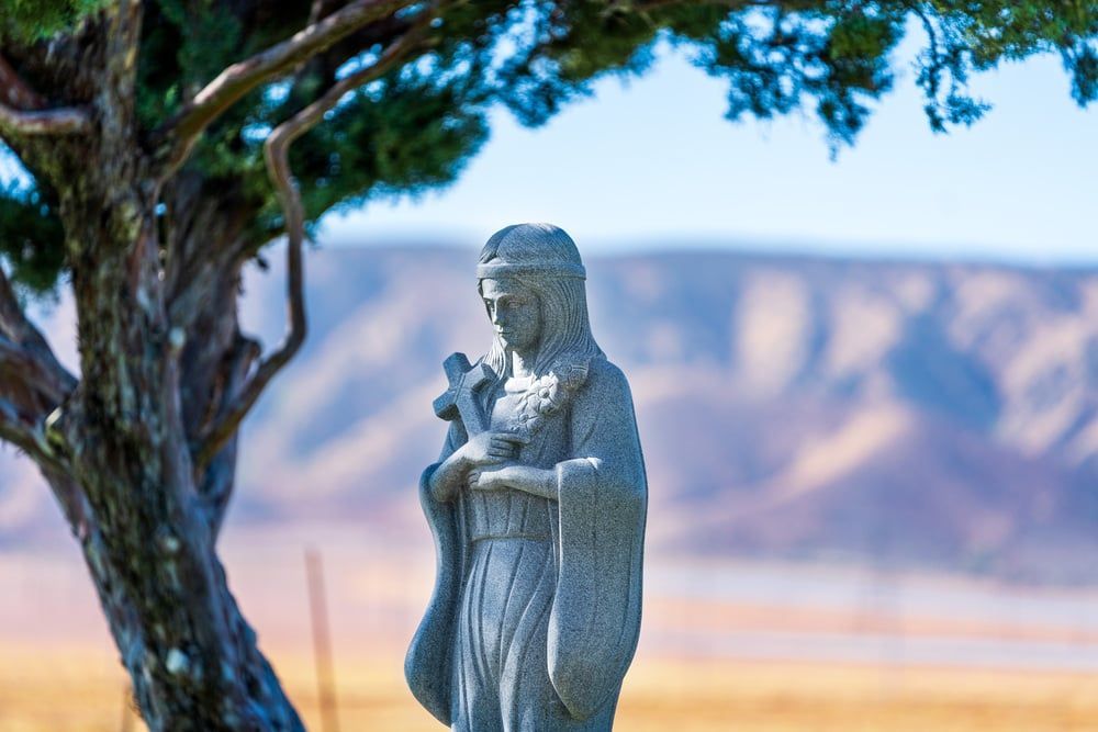 Stone statue of a robed figure beside a tree, with mountains and open landscape in the background.