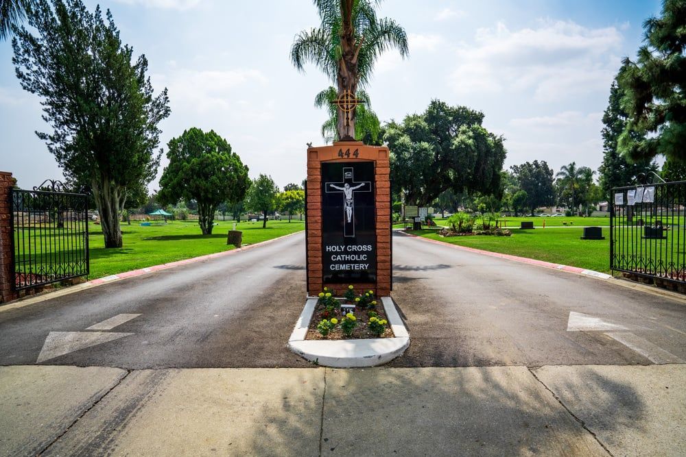 Gated driveway with a central sign monument, lined by palm trees and green lawns under a cloudy sky.