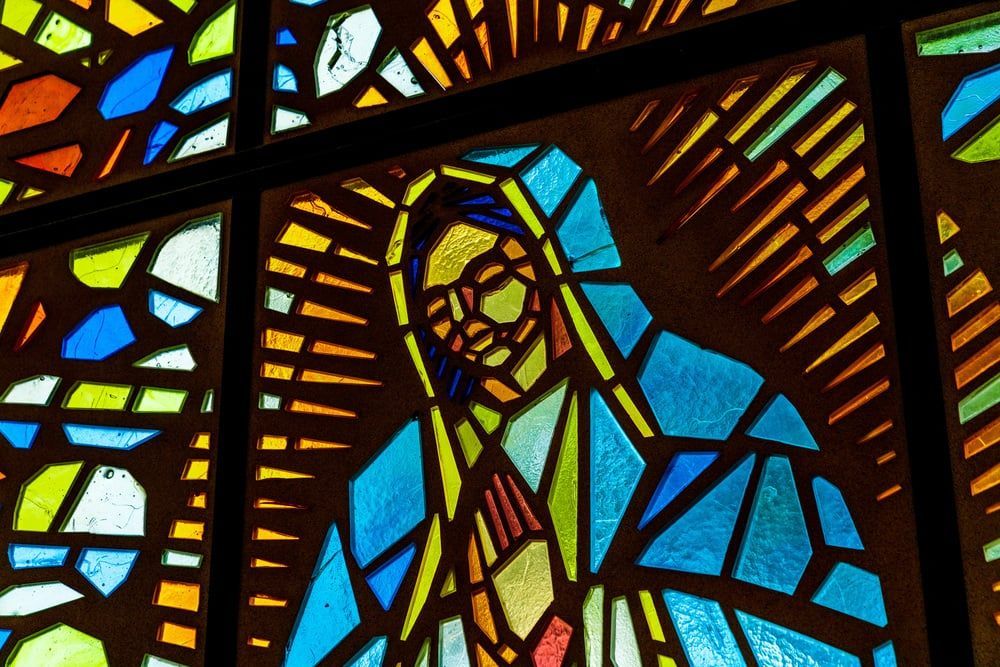 Colorful stained-glass window depicting a robed figure with a halo in blue, yellow, and red panels