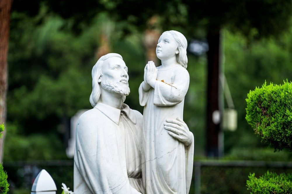 White statue of a kneeling man holding a child in a garden, with green trees and shrubs in the background