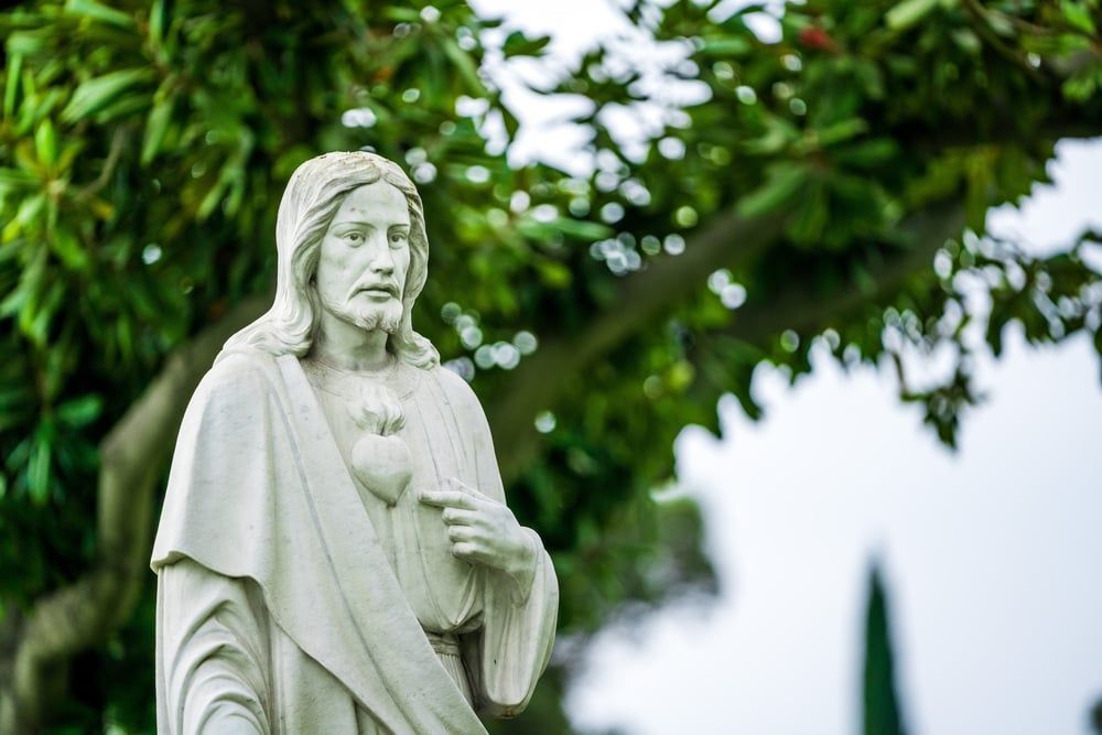 White stone statue of a robed figure outdoors, framed by green trees and foliage.