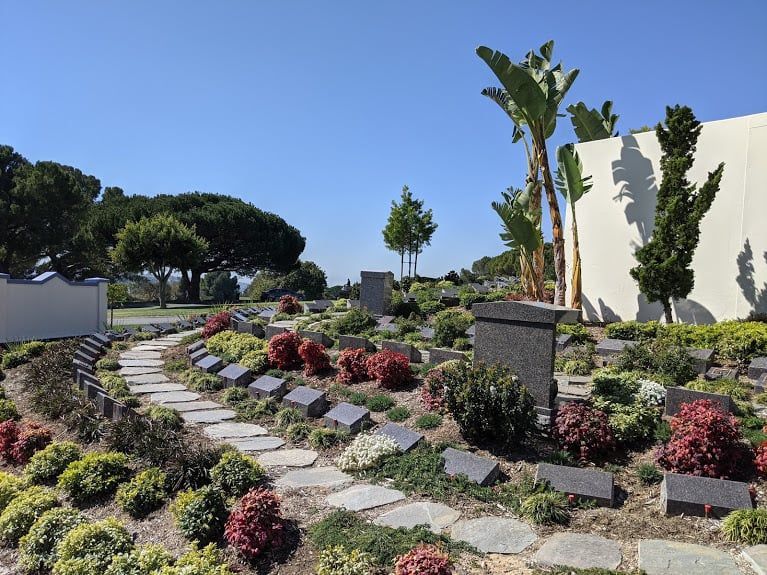 Curved stone path through a landscaped garden with red shrubs, banana trees, and white walls under a blue sky