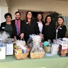 Six people stand behind gift baskets and boxes on a table at an indoor event.