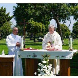 Two clergy at an outdoor altar during a religious service, surrounded by trees and flowers.
