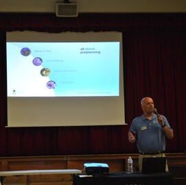 Man speaking into a microphone beside a projected slide in a dark lecture hall