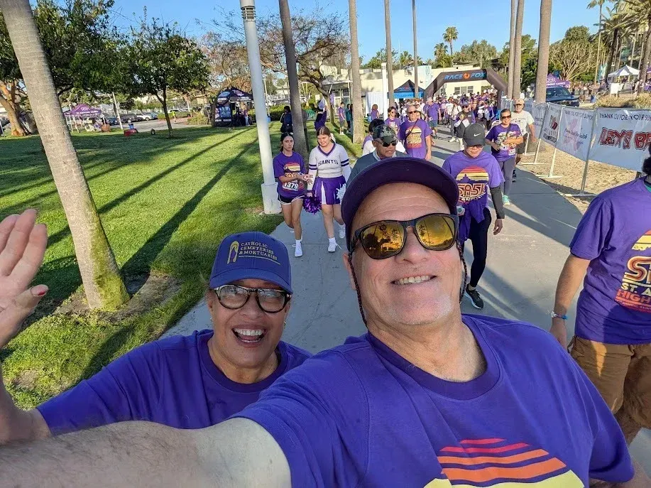 People in purple shirts walking outdoors on a sunny path, two smiling at the camera in the foreground