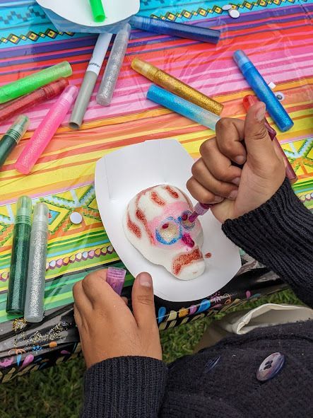 Hands painting a skull on paper with colorful markers on a striped tablecloth.