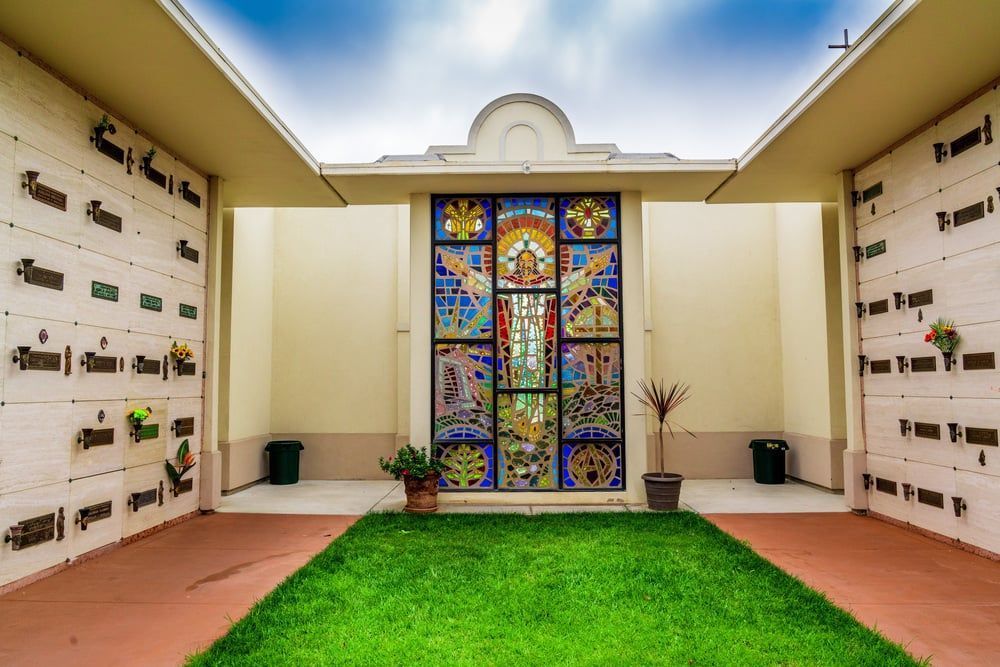 Courtyard with green lawn leading to a colorful stained-glass memorial wall and white surrounding niches