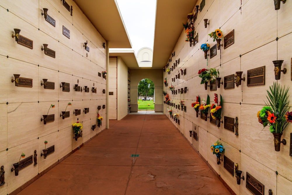 Narrow cemetery corridor with rows of wall niches and colorful flower bouquets.