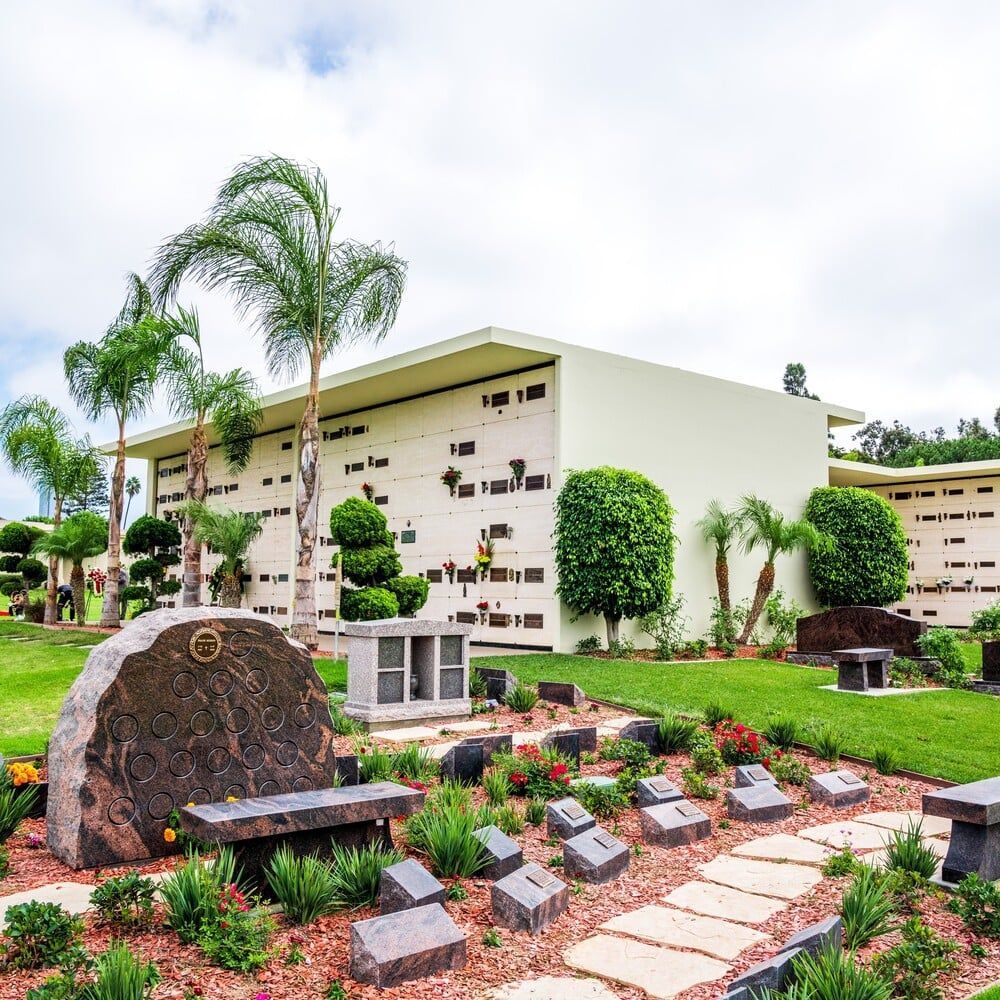Memorial garden with gravestones, benches, palm trees, and a white building under cloudy sky